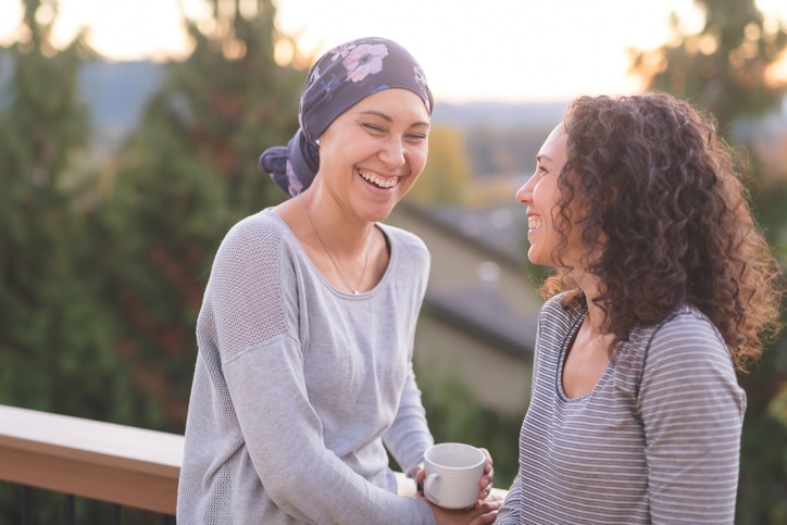 A beautiful young ethnic woman fighting cancer and wearing a head wrap talks with her sister on a porch. They are standing outdoors and there are mountains and trees in the background.