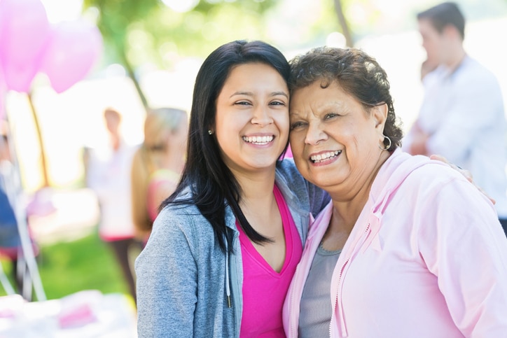 Senior Hispanic woman is smiling and looking at camera while hugging her young adult Hispanic granddaughter. Women are participating in breast cancer awareness race for charity. They are wearing pink athletic clothing. Other running teams are registering in background.
